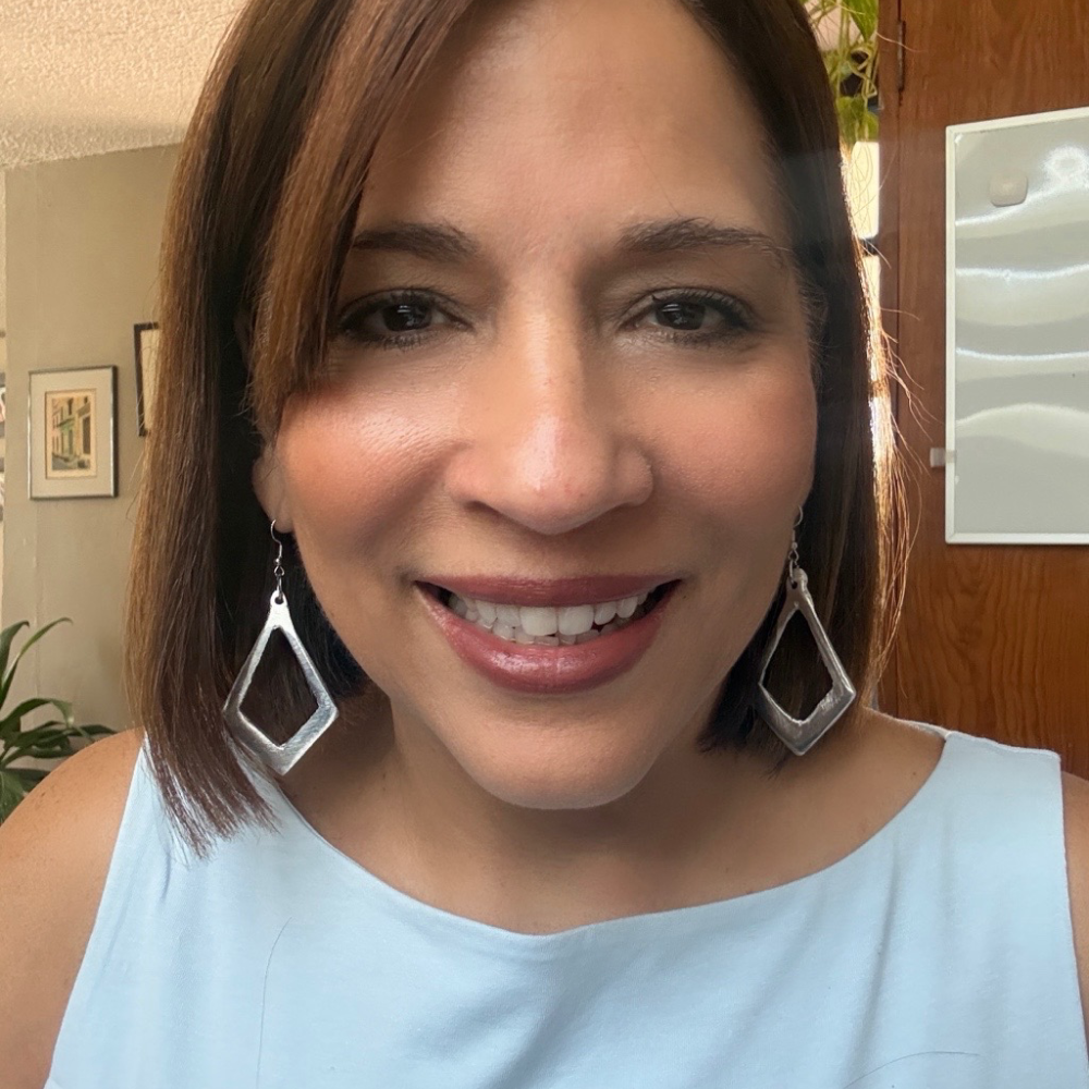 A smiling woman with shoulder-length brown hair wearing large silver geometric dangle earrings and a light blue sleeveless top, photographed indoors with a home background.